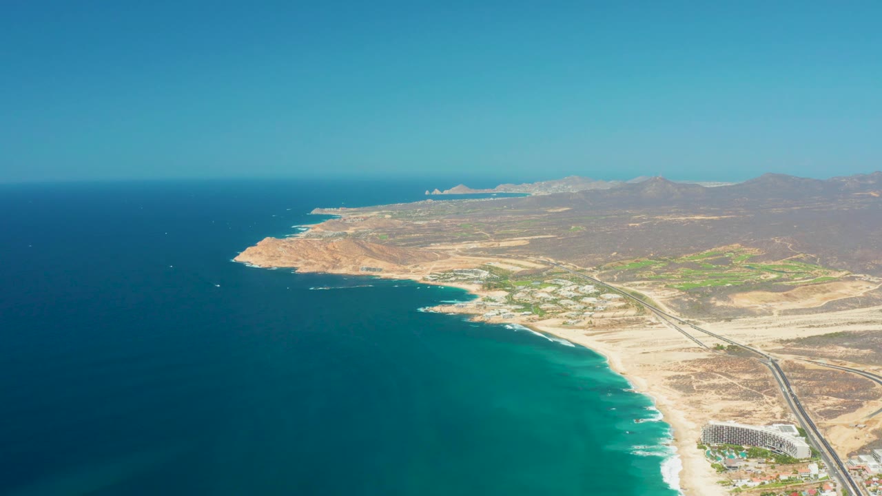 Aerial view of the Dreams Los Cabos coast with darker ocean offshore, pale sand along the right edge, resort-scale buildings near the lower right, dry inland terrain, and no visible people.