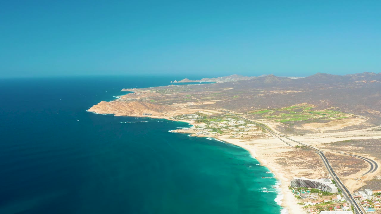 Aerial view at Dreams Los Cabos with the ocean on the left, a pale sandy shoreline, a road curving through the right side, scattered buildings, dry hills, distant mountains, and no visible people.