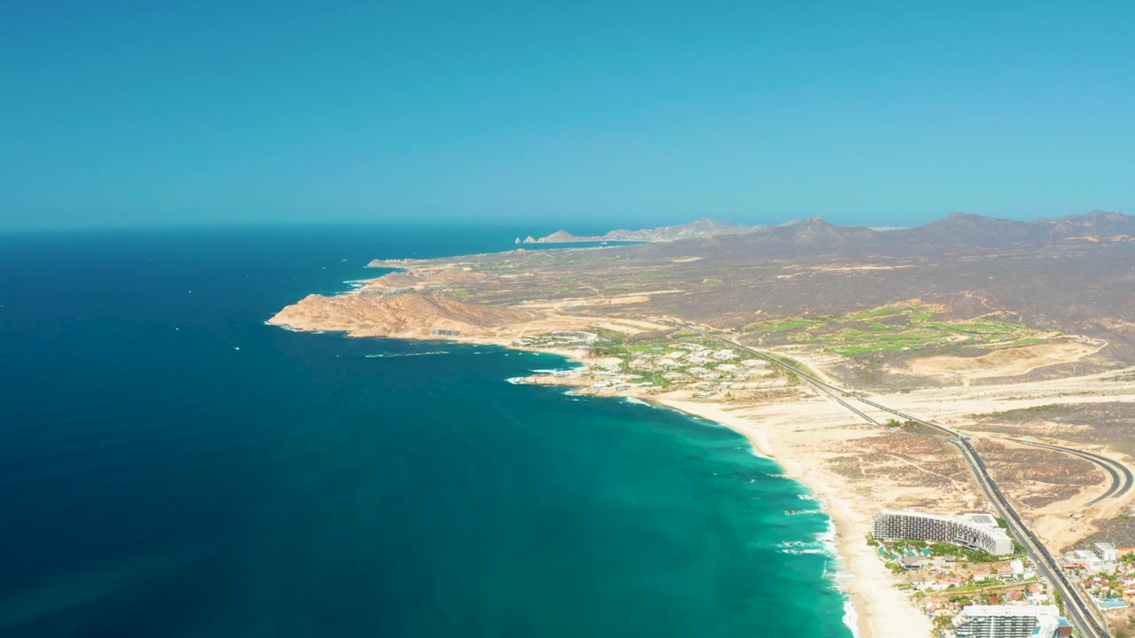 Aerial frame at Dreams Los Cabos showing the shoreline angled across the image, with open ocean on the left, beach and surf in the middle, a parallel road and beachfront buildings on the right, and no visible people.