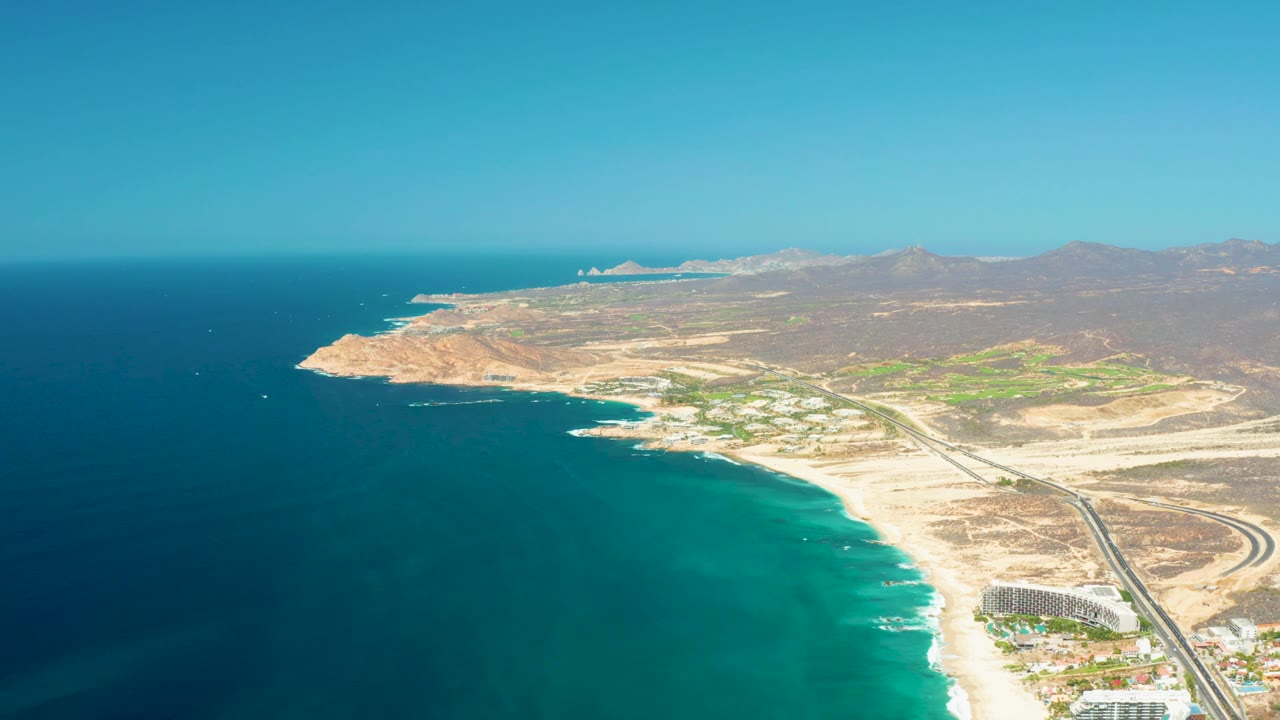 Mid-clip aerial view of the Dreams Los Cabos coast, with the ocean occupying the left foreground, a bright sandy beach and surf line below, a road and buildings to the right, hazy hills inland, and no visible people.