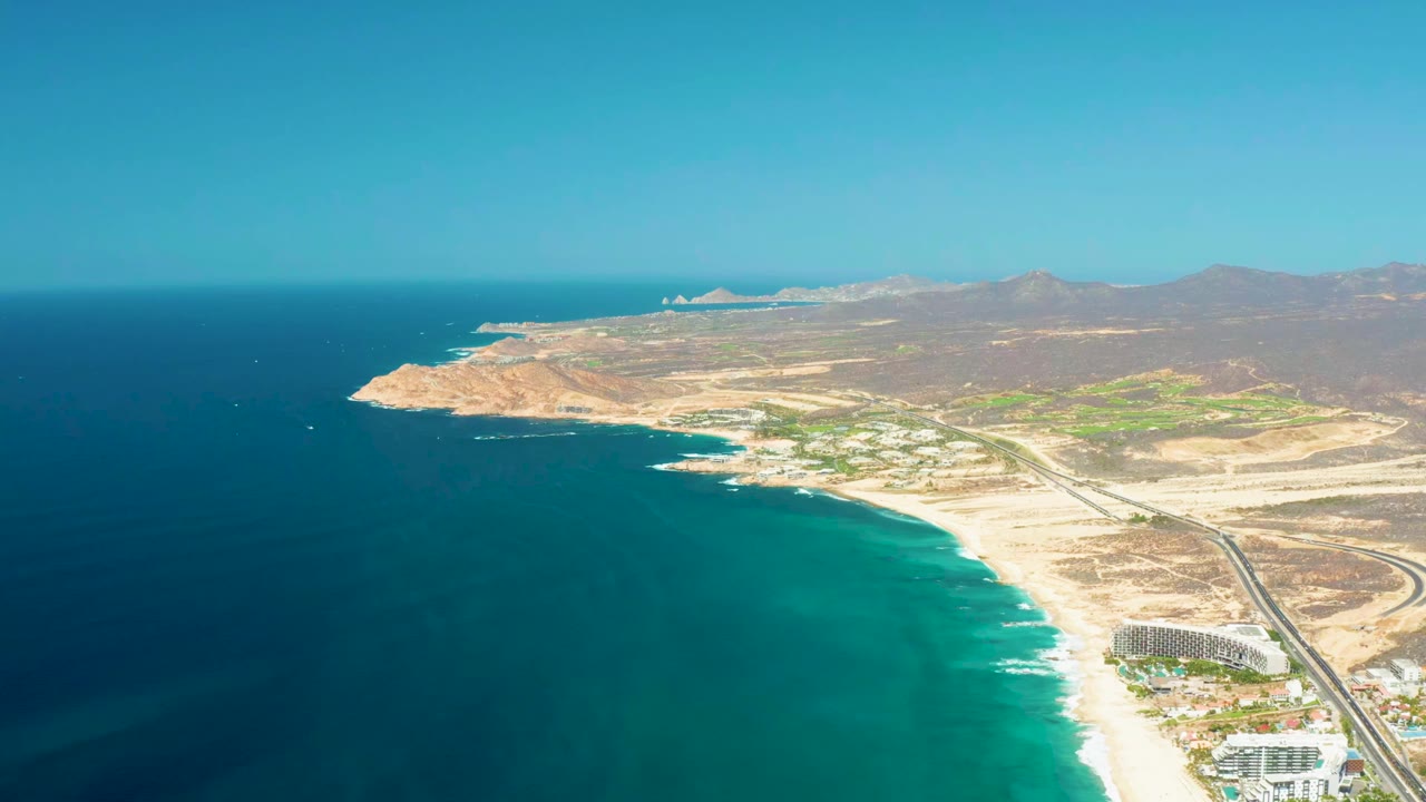 High aerial frame over Dreams Los Cabos showing deep blue and turquoise water, pale sand, a road parallel to the shore, small beachfront structures, distant arid hills, and no visible people.