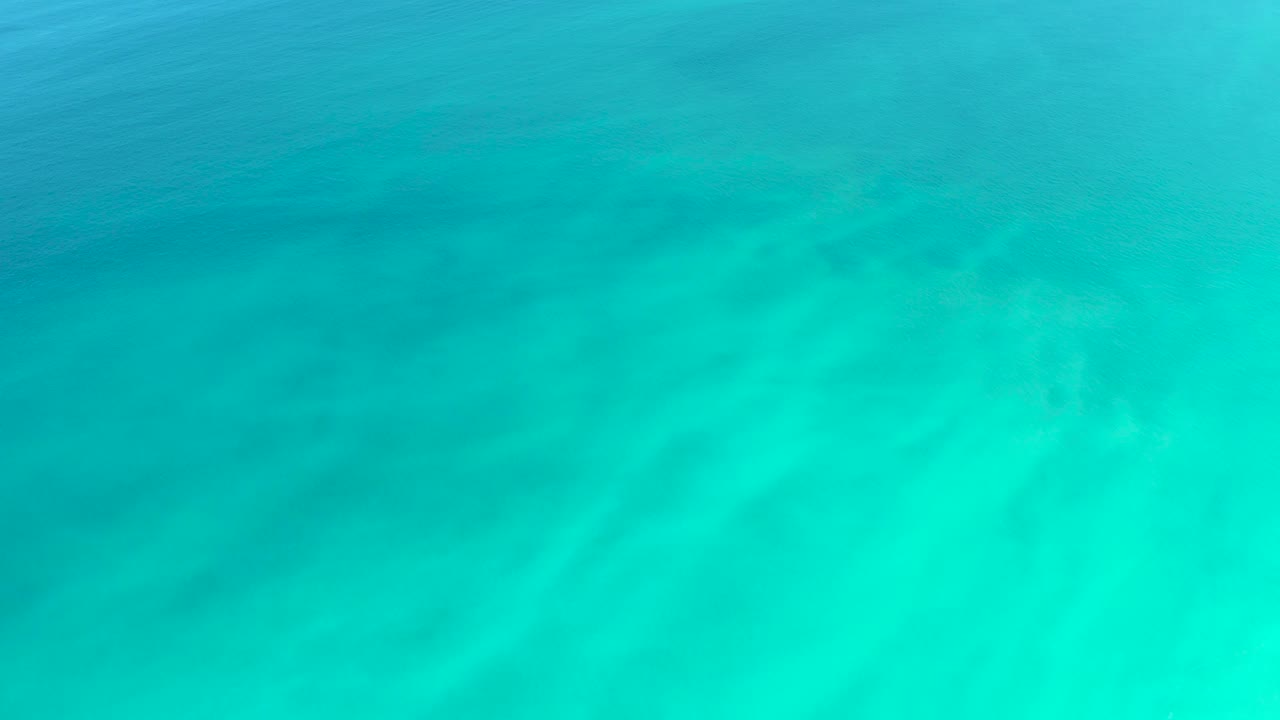 Bright turquoise shallow water at Dreams Los Cabos reveals pale sand beneath the surface, with soft wave texture and no visible people.