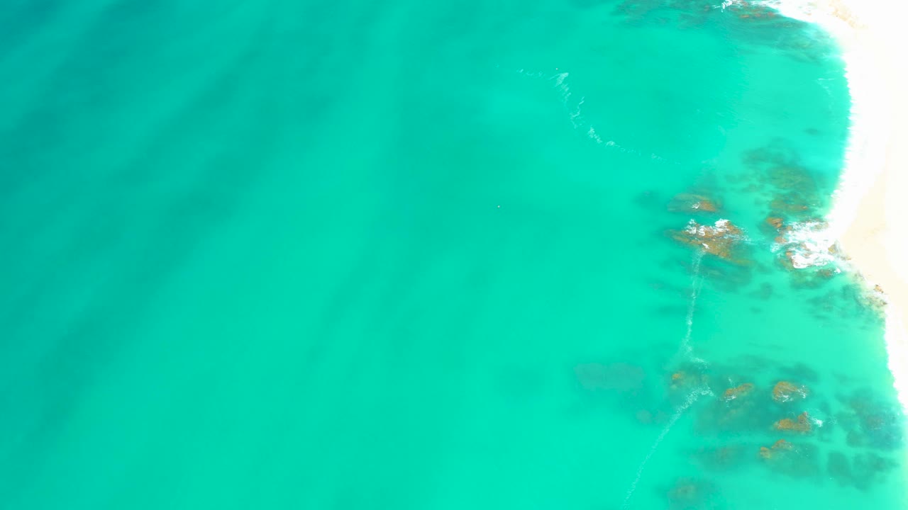 Aerial frame from Dreams Los Cabos showing clear turquoise water, a bright strip of sand on the right, small white wave wash, submerged rocks, and no visible people.