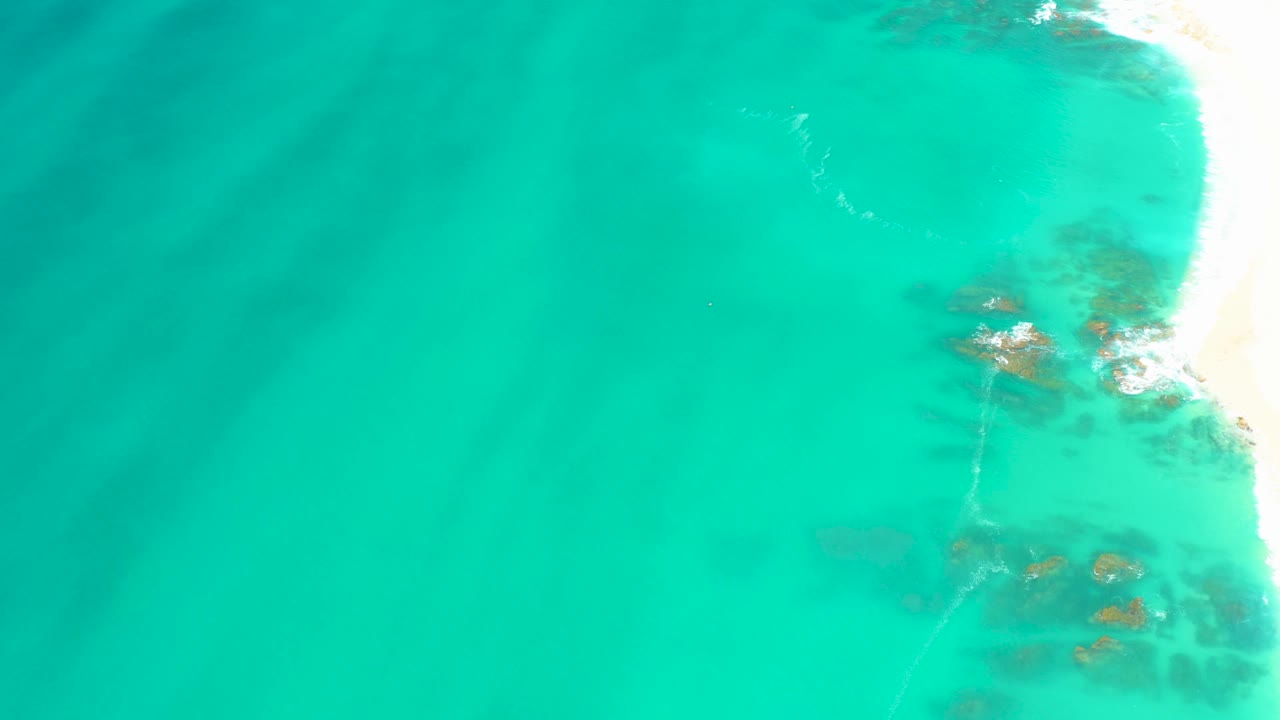 High aerial view at Dreams Los Cabos with open turquoise water across most of the frame, bright sandy shoreline at right, faint reef shapes below, and no visible people.