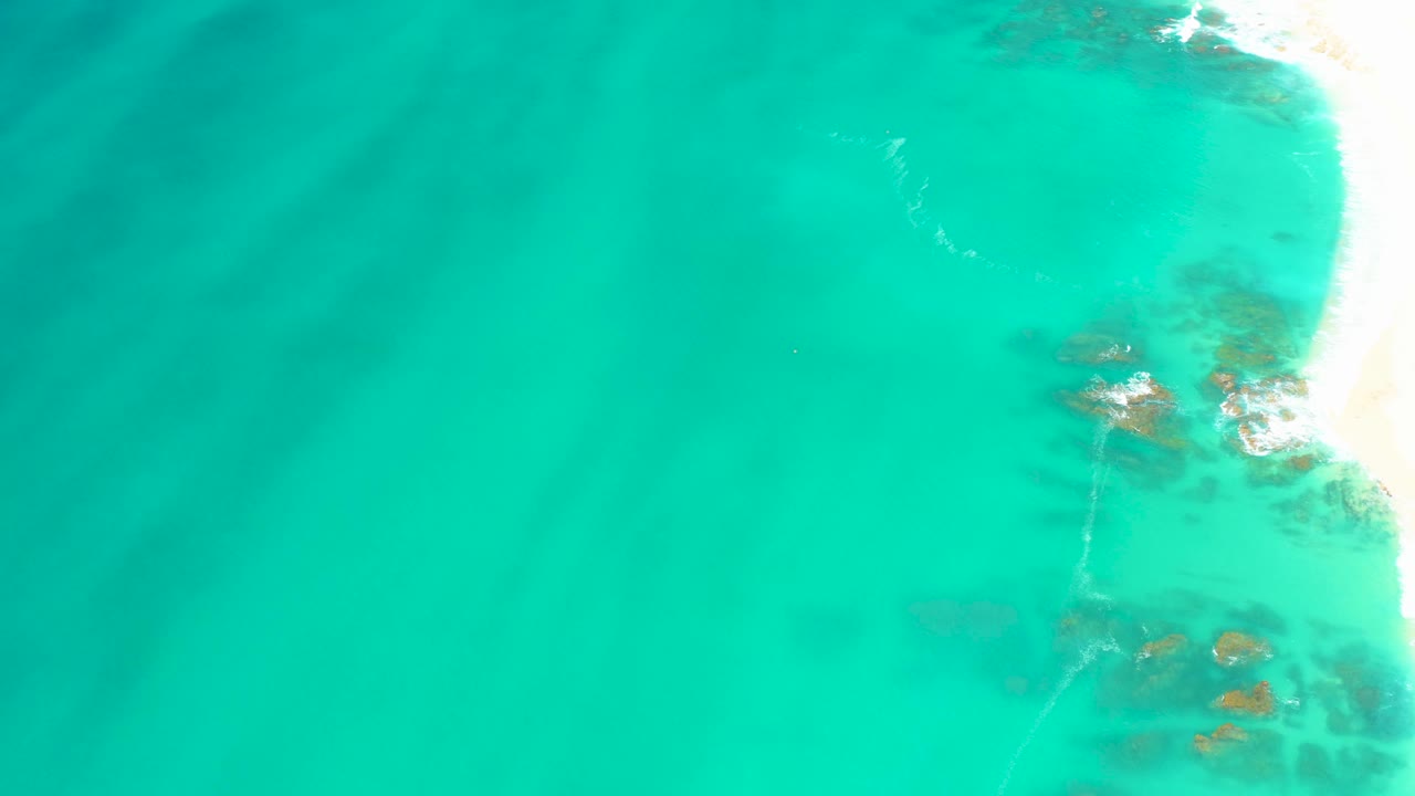 Turquoise sea over sand with darker underwater patches and surf at the right edge.