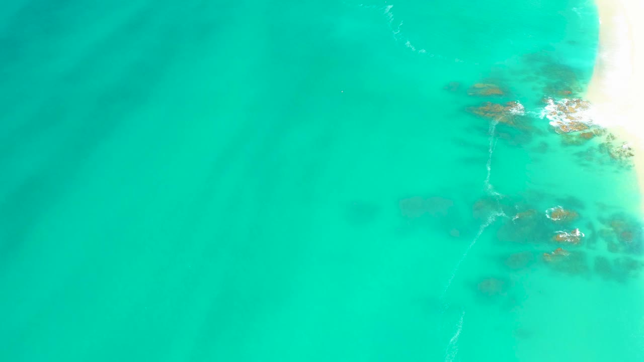 Aerial frame from Dreams Los Cabos showing blue-green water, faint seabed shapes, clustered rocks along the right side, foam at the shoreline, and no visible people.