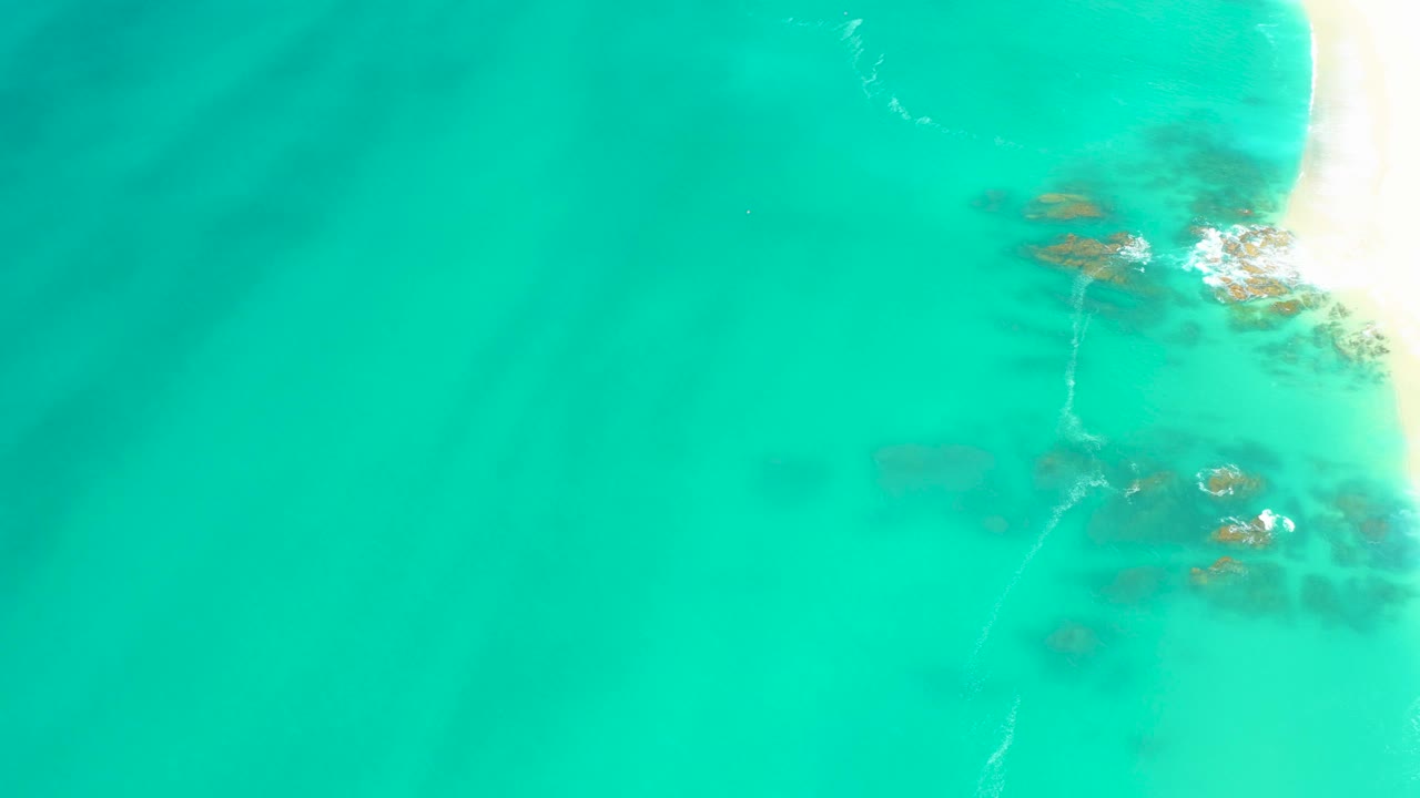 Overhead Dreams Los Cabos view with broad turquoise shallows, darker underwater rocks at the right, a narrow pale beach edge, white foam, and no visible people.