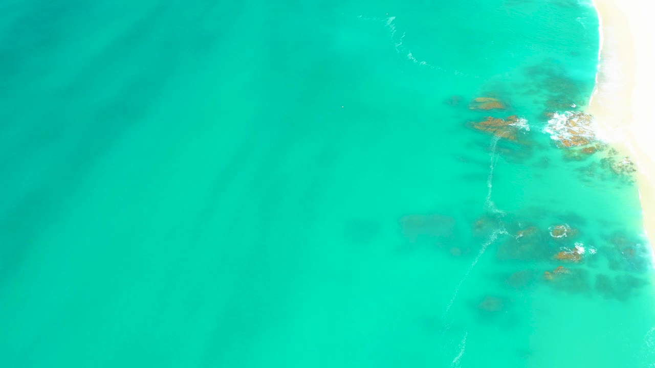 Mid-clip aerial view at Dreams Los Cabos with the beach edge brighter on the right, rocks visible beneath clear water, light foam lines, and no visible people.