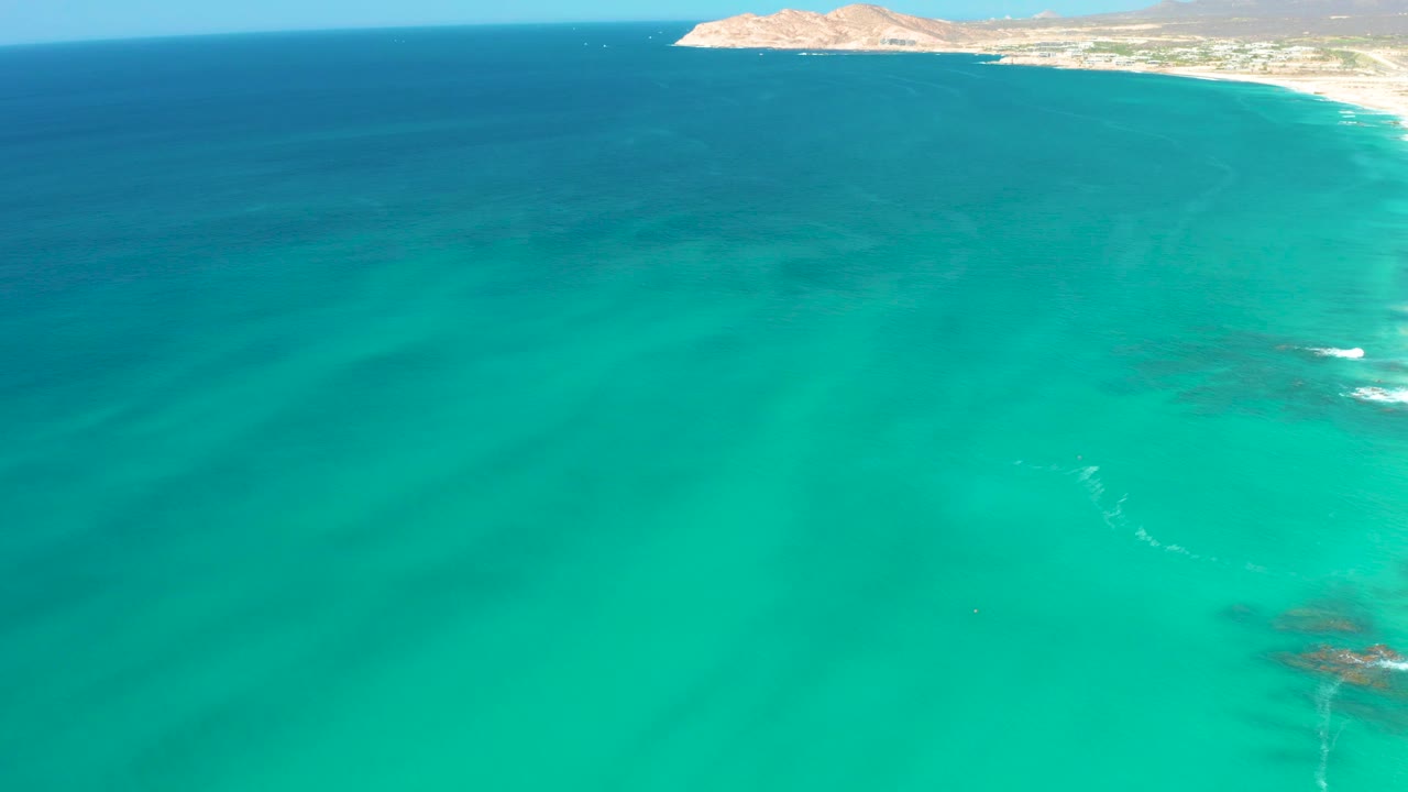 Aerial view at Dreams Los Cabos with clear blue-green water dominating the frame while the shoreline and hill remain narrow along the top, no visible people.