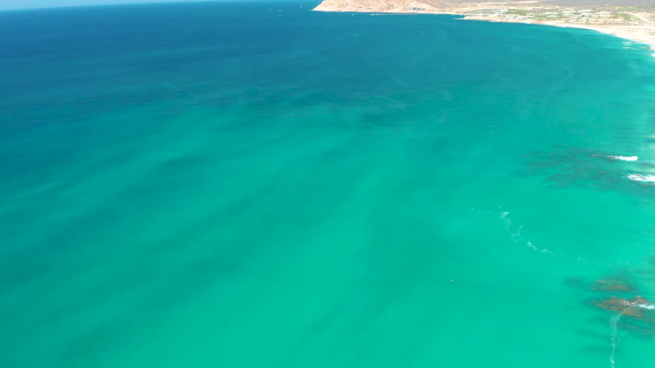 Aerial frame over blue-green water at Dreams Los Cabos, with the coastline reduced to a thin top-right strip and no visible people.