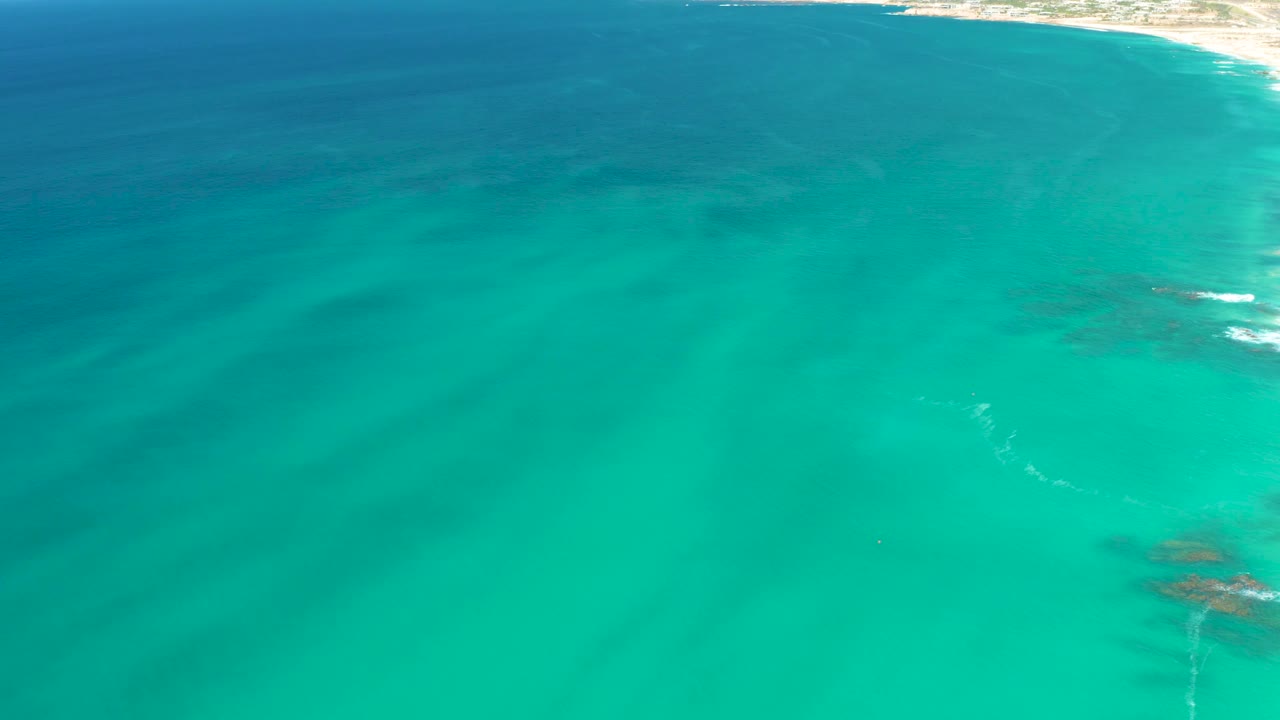 Aerial view at Dreams Los Cabos showing mostly turquoise ocean, darker submerged patches, and a small remaining shoreline area near the upper right, no visible people.