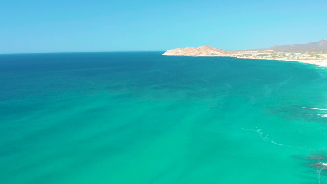 Opening aerial view at Dreams Los Cabos, Mexico, showing turquoise ocean in the foreground, a sandy shoreline and rocky headland near the horizon, and no visible people.