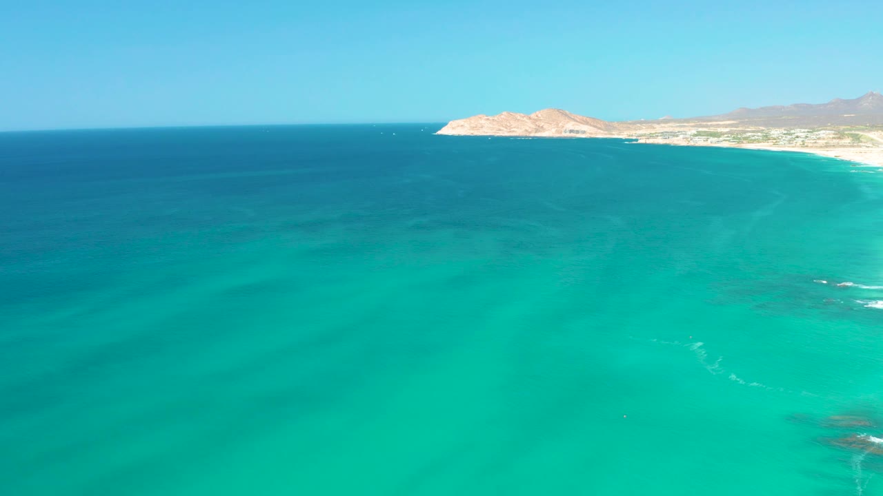 Aerial view near Dreams Los Cabos with clear turquoise water across the frame, small white wave streaks over shallow rocks, the beach at upper right, and no visible people.