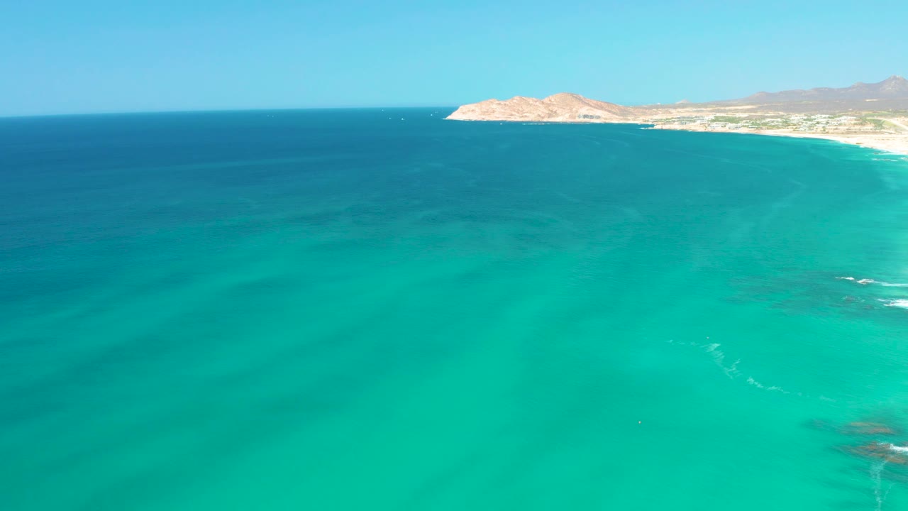 Mid-clip aerial frame at Dreams Los Cabos showing the turquoise bay, darker blue water near the horizon, sandy coast at the upper right edge, rocky headland beyond it, and no visible people.