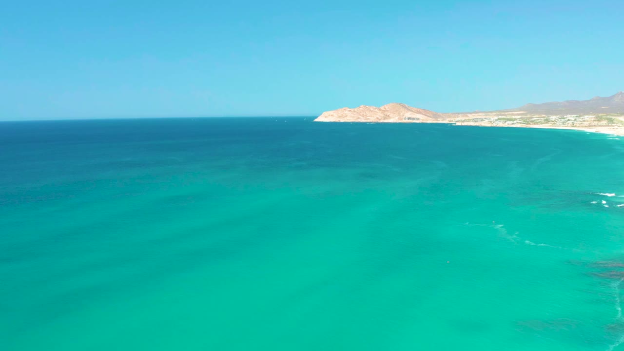 A second aerial frame holds on open turquoise sea at Dreams Los Cabos, with the pale beach and rocky headland running across the upper edge and no visible people.