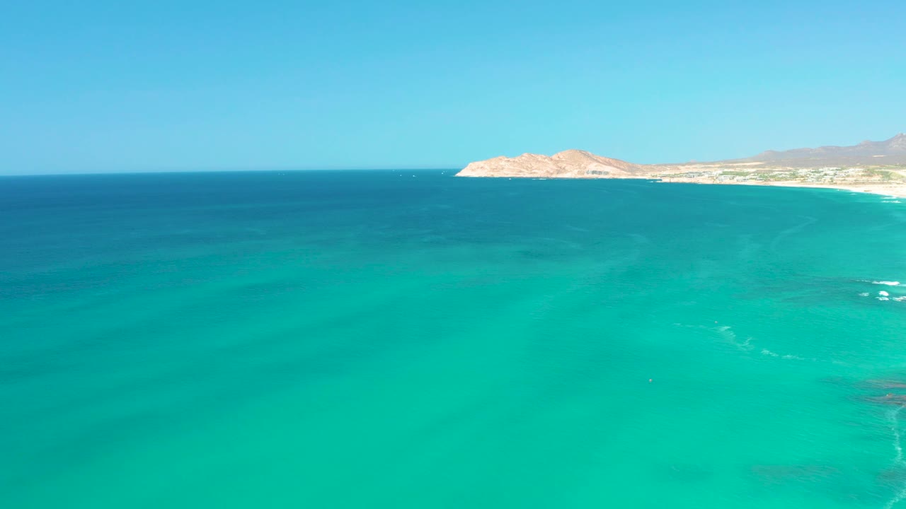 The aerial view continues over clear turquoise water near Dreams Los Cabos, with faint white surf marks below, the coastline high in frame, and no visible people.