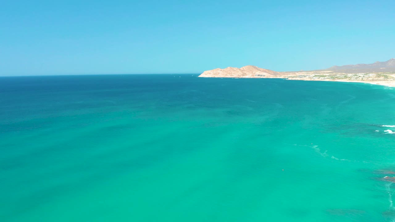 Mid-clip aerial frame at Dreams Los Cabos showing turquoise ocean across the foreground, small wave foam near shore, a rocky headland, distant hills, and no visible people.