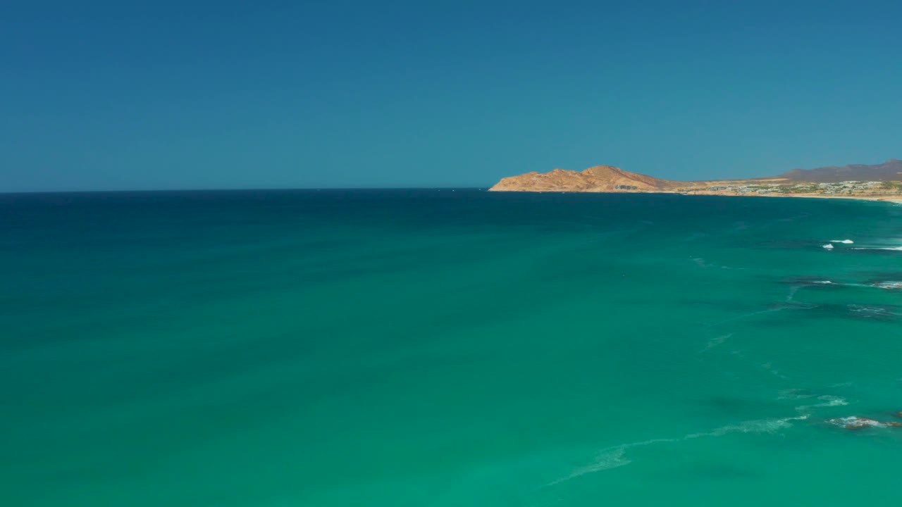 Clear turquoise water stretches toward the horizon at Dreams Los Cabos, with small whitecaps and a sandy rocky coast in the background, and no visible people.
