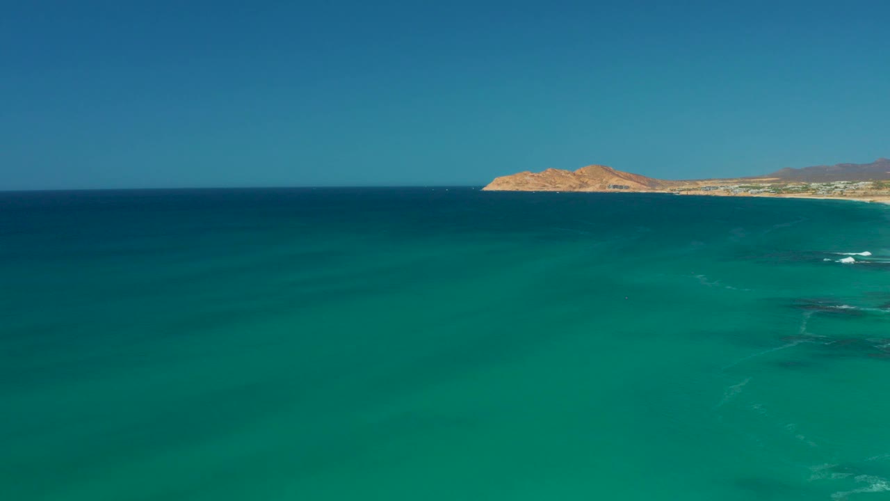 A wide view of turquoise sea leads to a sandy coastline and low rocky landforms near Dreams Los Cabos, with small white wave marks and no visible people.
