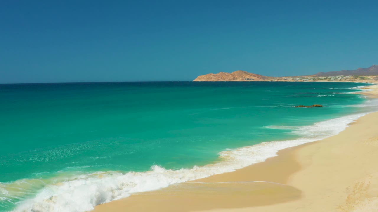 Turquoise ocean waves breaking beside a sandy beach under a clear sky.