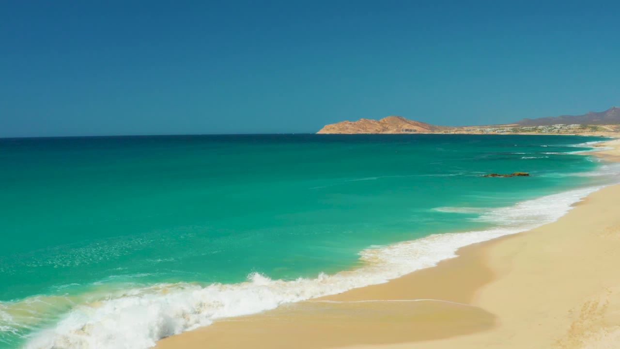 Dreams Los Cabos beach at 0:01 with a wider band of white foam crossing the lower sand, clear turquoise water, distant land, and no visible people.