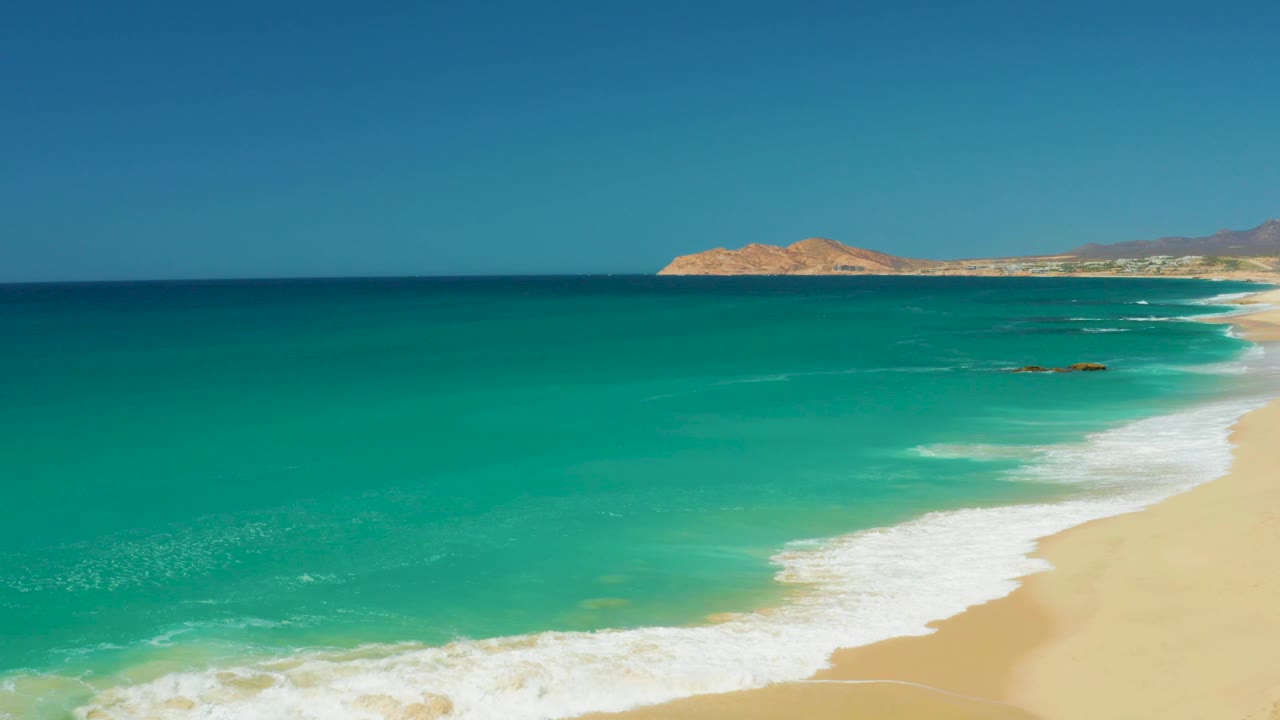 Dreams Los Cabos shoreline at 0:02 with the beach pushed to the right edge, a curling white surf band, open turquoise water, and no visible people.