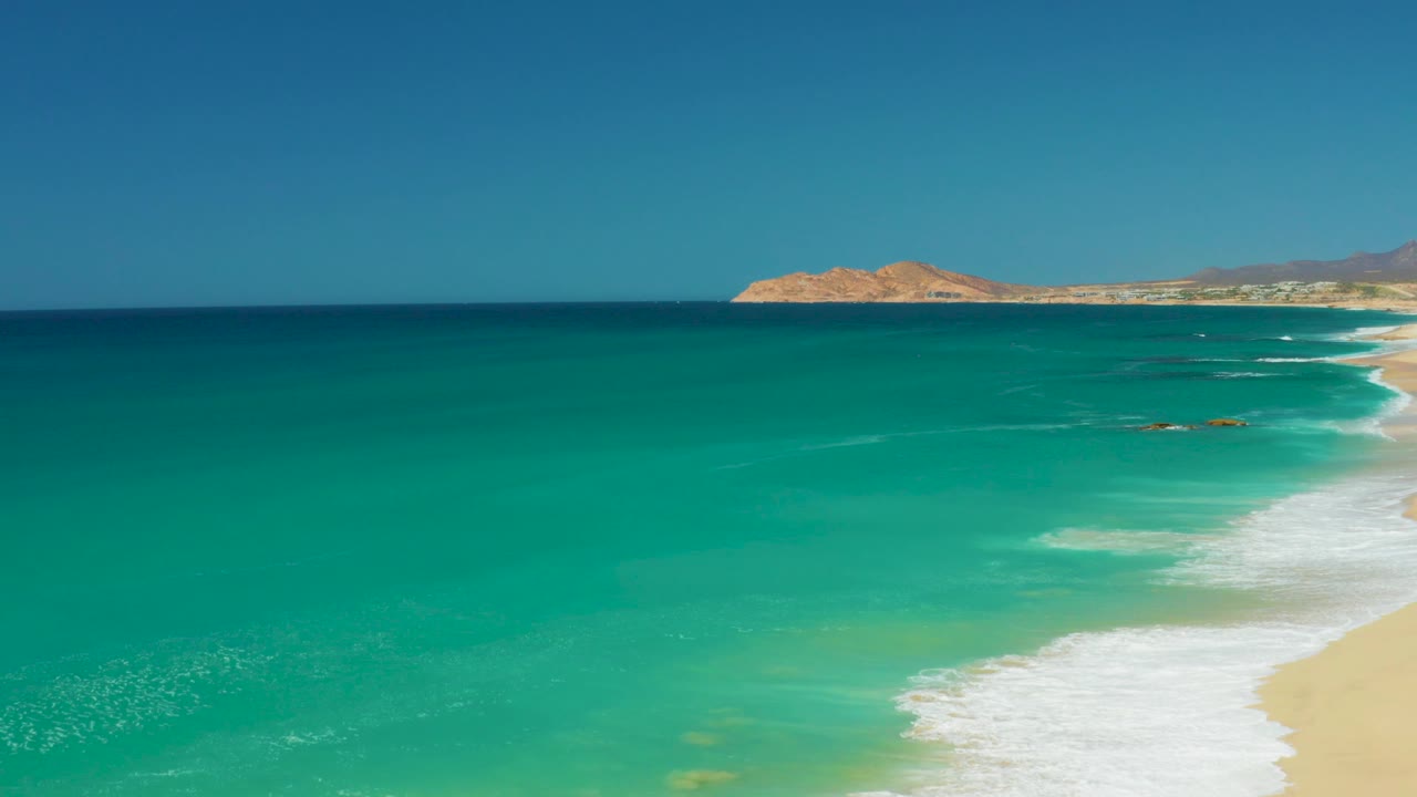 Dreams Los Cabos aerial view at 0:03 where open water fills most of the frame, the beach narrows on the right, and no visible people appear.