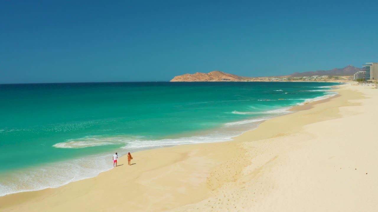 Aerial view at Dreams Los Cabos, Baja California Sur, with two people on pale sand near turquoise water and low hills on the horizon.
