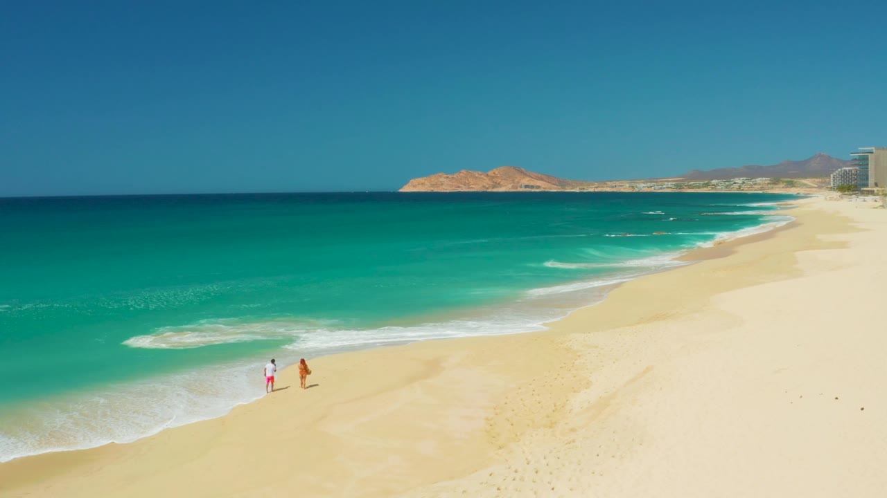 Two walkers on open sand near white foam and clear blue water.