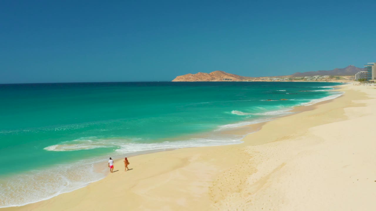 Aerial frame of the Dreams Los Cabos shoreline with two people closer to the breaking wave line and tan hills beyond the turquoise sea.