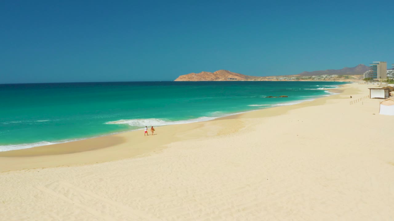 At Dreams Los Cabos in Baja California Sur, two small figures walk near the waterline as turquoise surf curves across a broad pale beach under a clear blue sky.