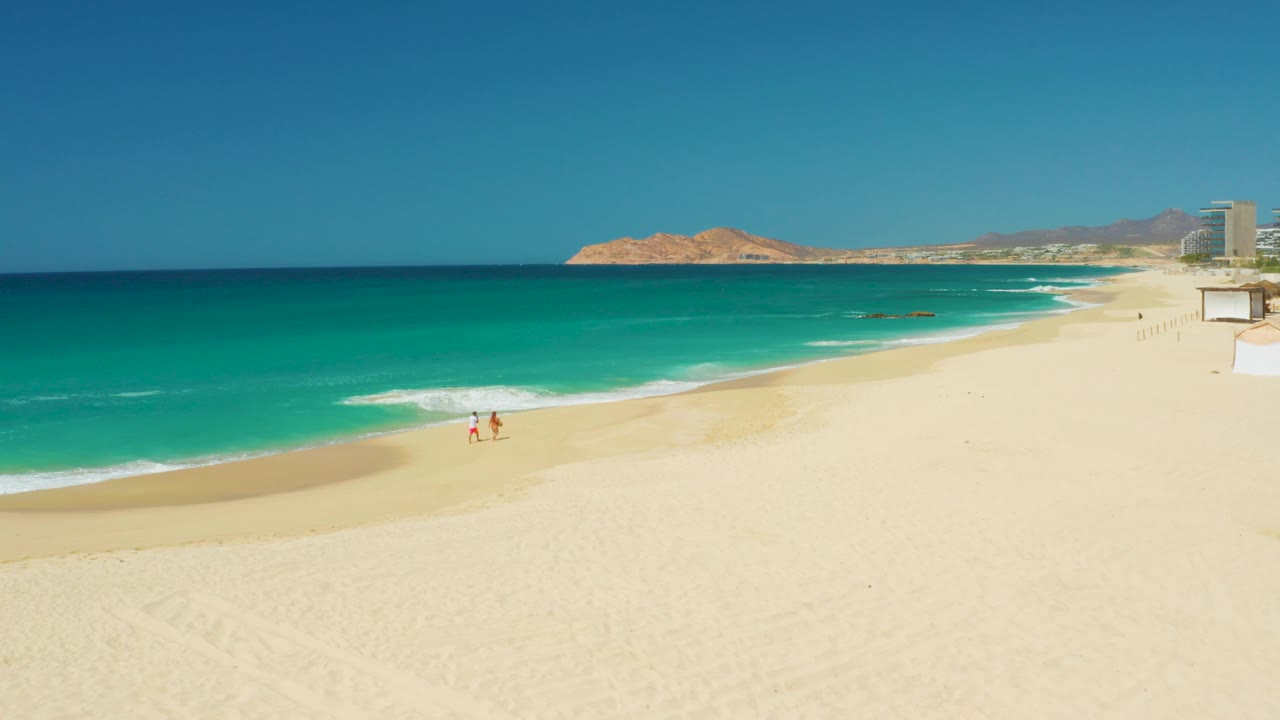 Two walkers on pale sand with turquoise water and distant rocky shoreline.