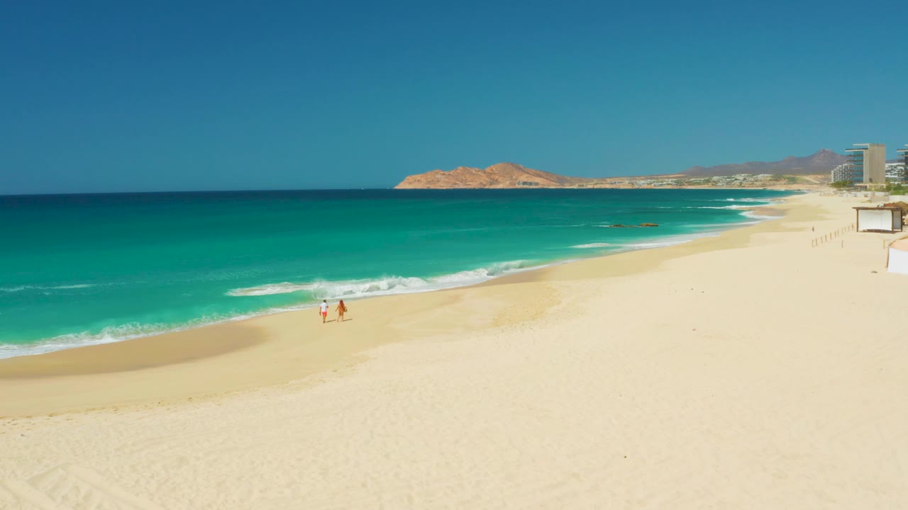 At 0:02, the two walkers appear slightly farther along the water's edge, with more white foam visible near them and the rocky horizon running behind the open beach.