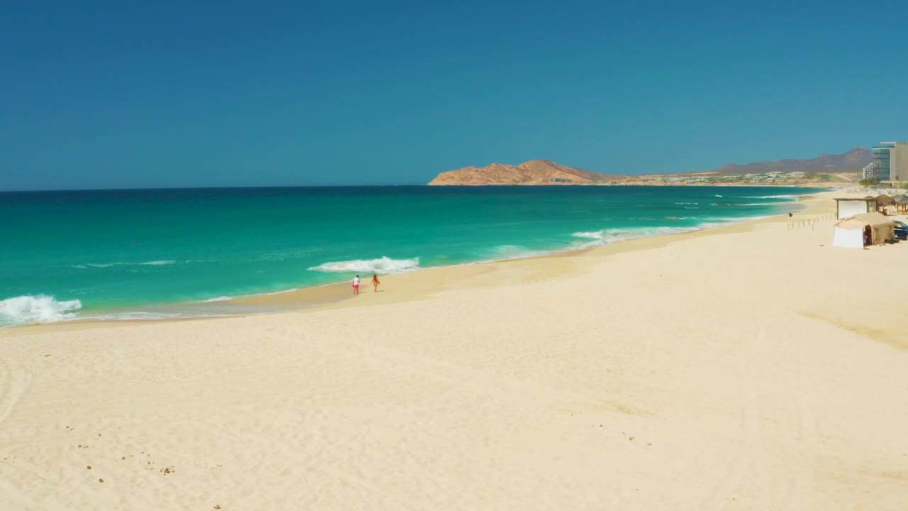 Wide Dreams Los Cabos beach view with a curved surf edge, two distant people walking near the wet sand, clear sky, hills, and resort structures at the far right.