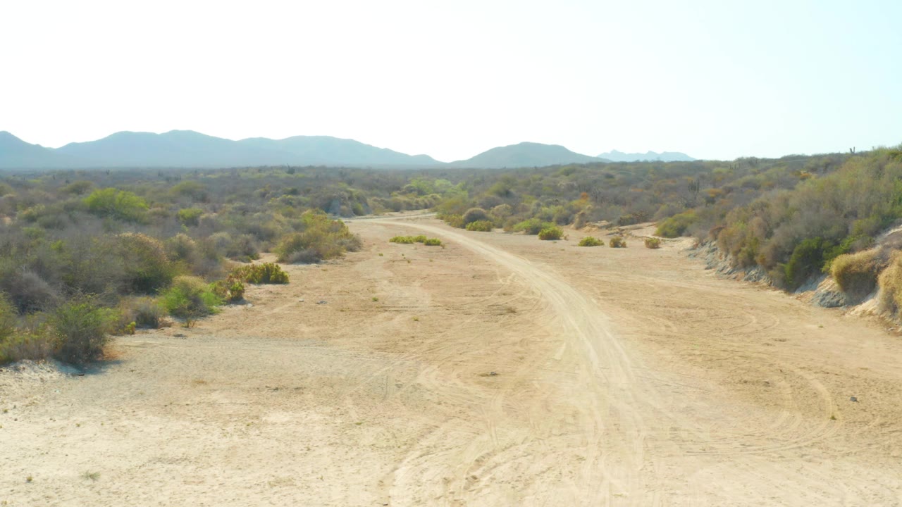 Opening frame of a sandy dirt road in Los Frailes, Los Cabos, Baja California Sur, with low scrub on both sides, distant mountains, clear bright sky, and no visible people.