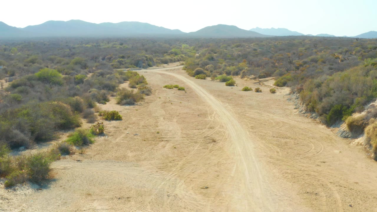 The sandy road sits slightly nearer in the frame as it crosses sparse Los Frailes bushland toward hazy low mountains, with no visible people or vehicles.