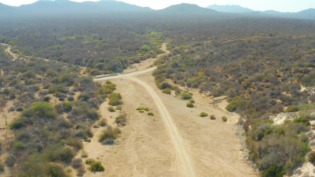 Aerial view of a pale dirt road curving through dry scrubland toward distant mountains.