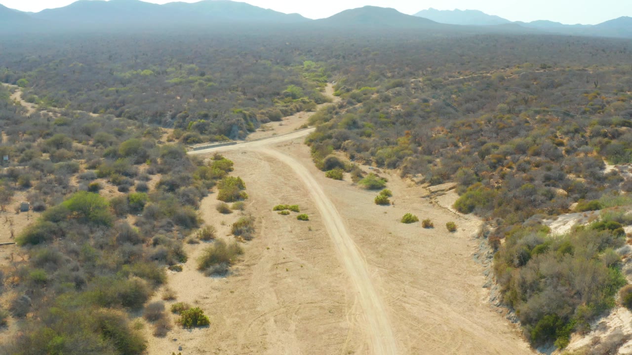 Aerial view over Los Frailes, Los Cabos, Baja California Sur, with the winding dirt track more centered, brush-covered ground spreading across the frame, hazy mountains in back, and no visible people.