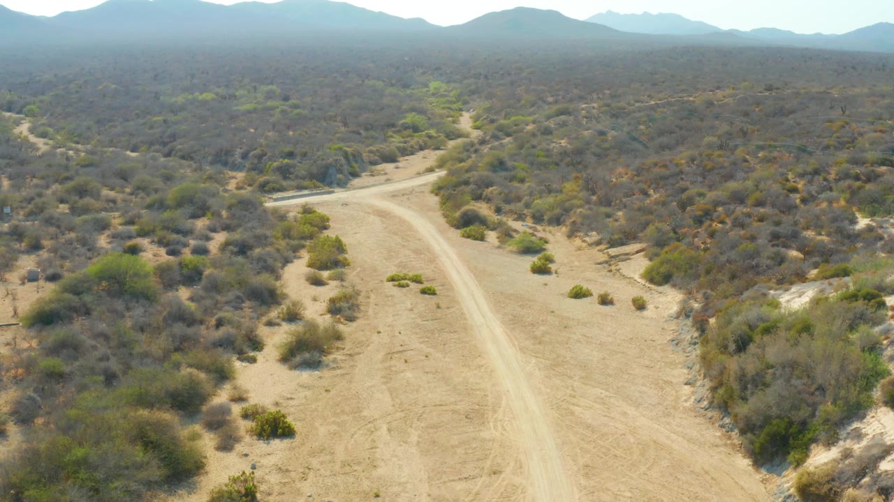Mid-clip aerial frame in Los Frailes, Los Cabos, Baja California Sur, showing the main dirt road through dry scrub with a pale side track or crossing visible ahead and no visible people.