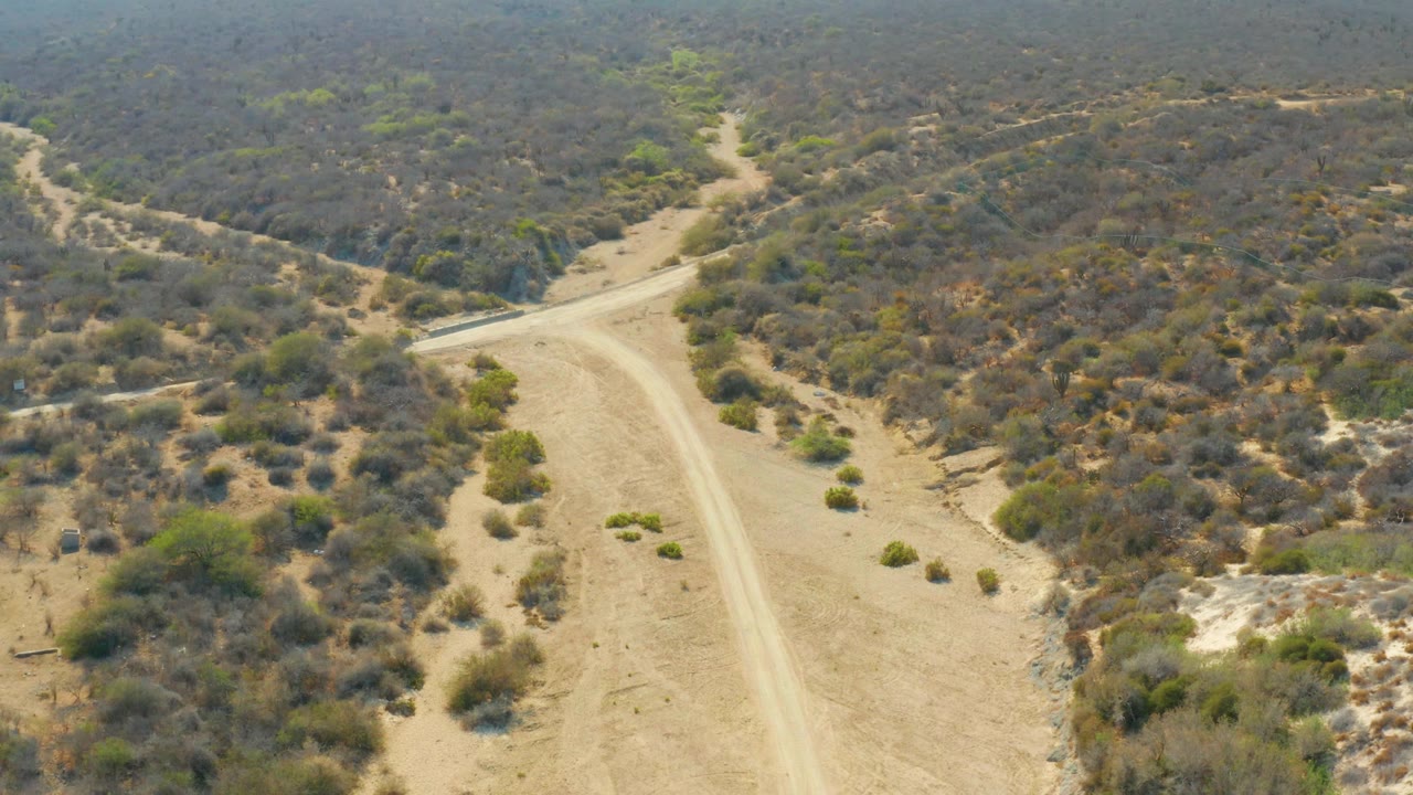 Elevated view over Los Frailes dry terrain, with the sandy road bending past scrub-covered slopes and small green bushes, while distant hills sit in haze and no people are visible.