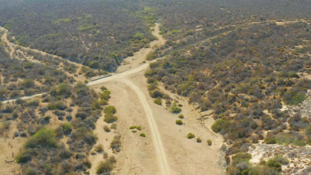 Elevated frame from Los Frailes showing the main dirt road curving through sandy ground, with scrub-covered hills and no visible people.
