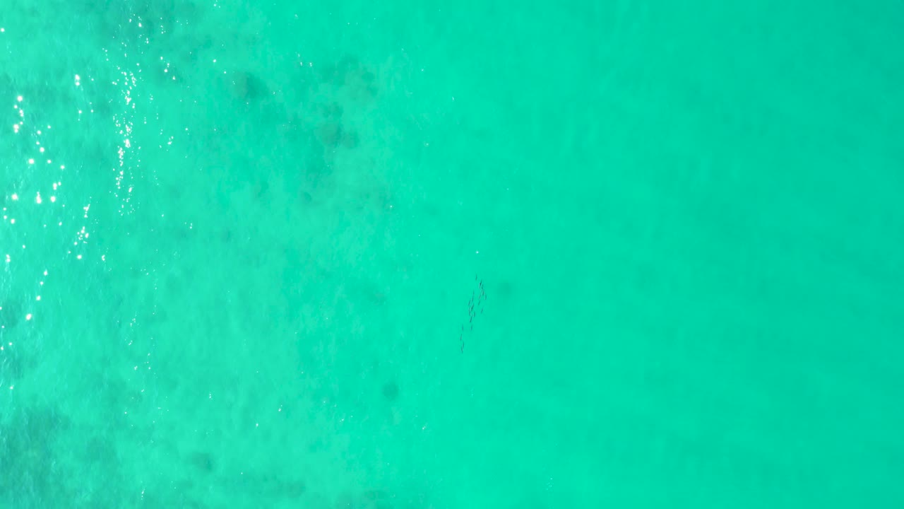 Aerial view over shallow turquoise ocean at Los Frailes, showing scattered darker seabed patches and a vertical band of sparkling surface light, with no visible people in this still.
