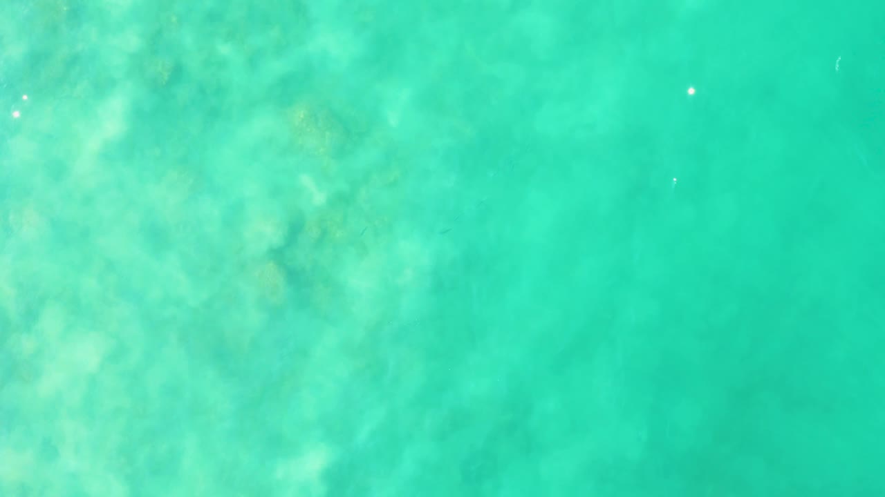 Top-down frame of Los Frailes turquoise sea with cloudy underwater formations and a few small bright reflections on the surface, no visible people.