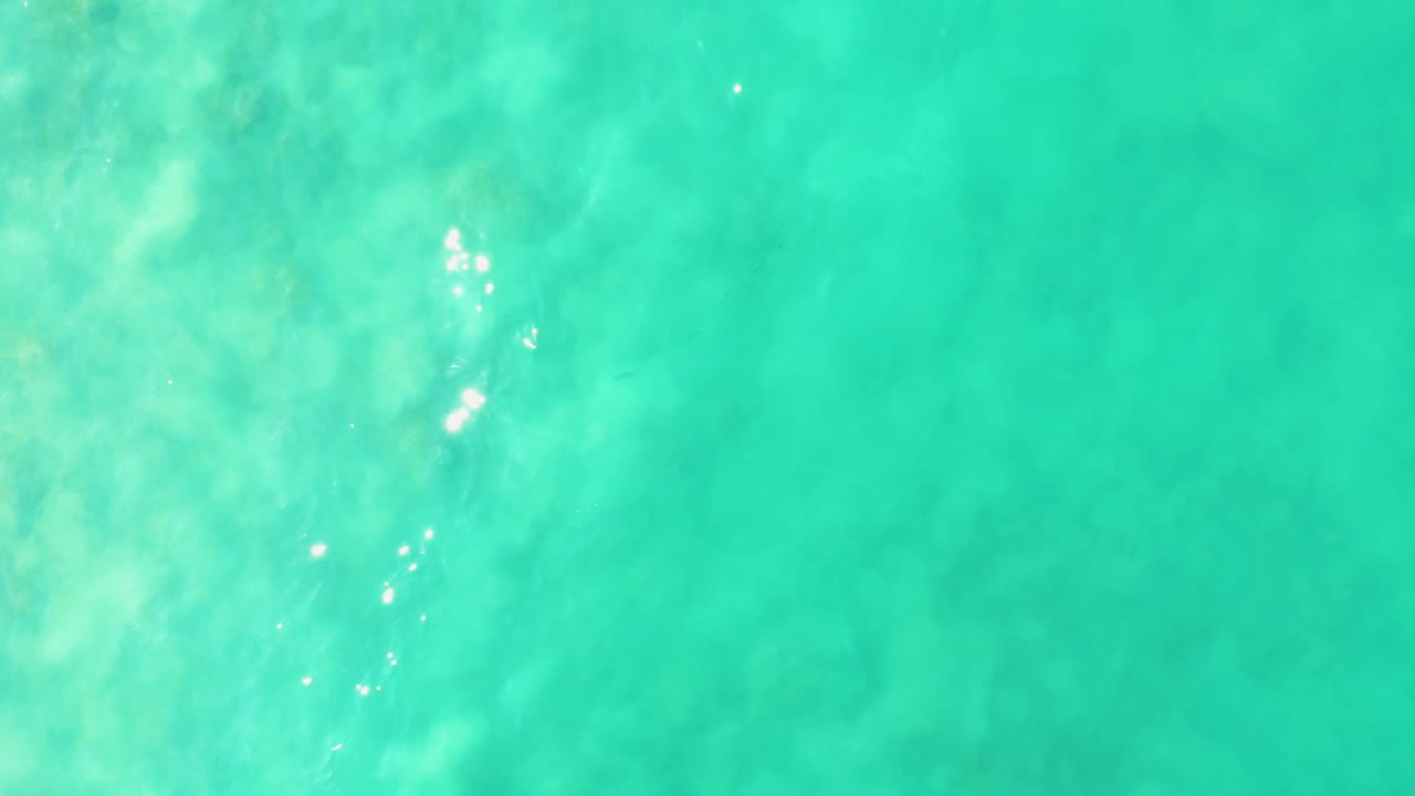 Clear Los Frailes ocean water seen from above, with a brighter cluster of sun sparkle over pale submerged rocks or coral and no visible people.