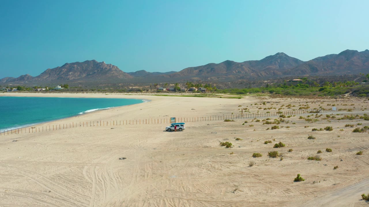 Los Frailes beach appears from above with blue water along the left edge, broad open sand, a small parked vehicle, sparse desert plants, and no visible people.