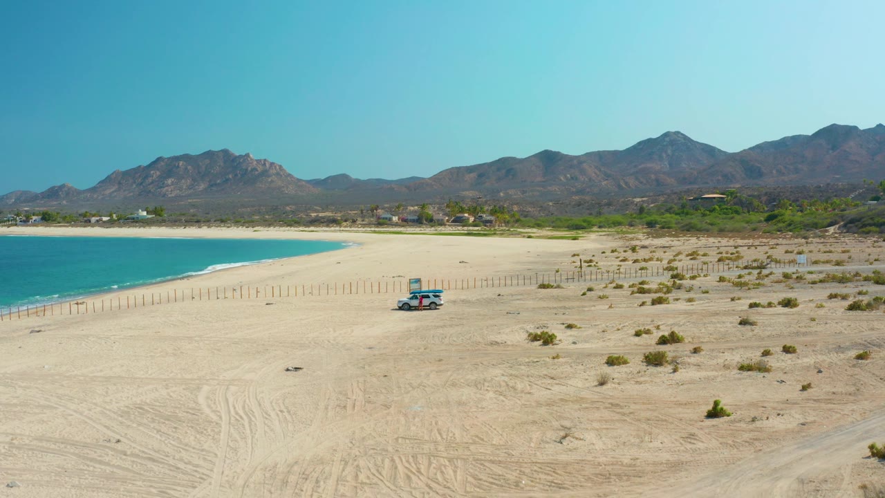 Wide coastal desert beach with vehicle, fence posts, turquoise water, and mountains under clear sky.
