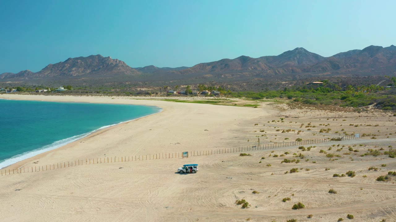Small vehicle on a sandy beach beside blue ocean and mountains.