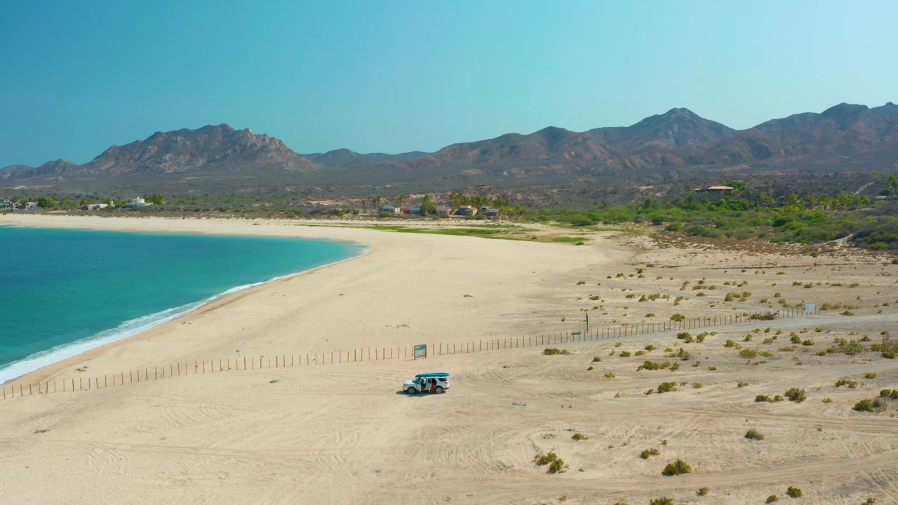 Los Frailes beach scene with the small vehicle slightly farther right on the sand, turquoise water curving along the left edge, distant mountains, and no visible people.