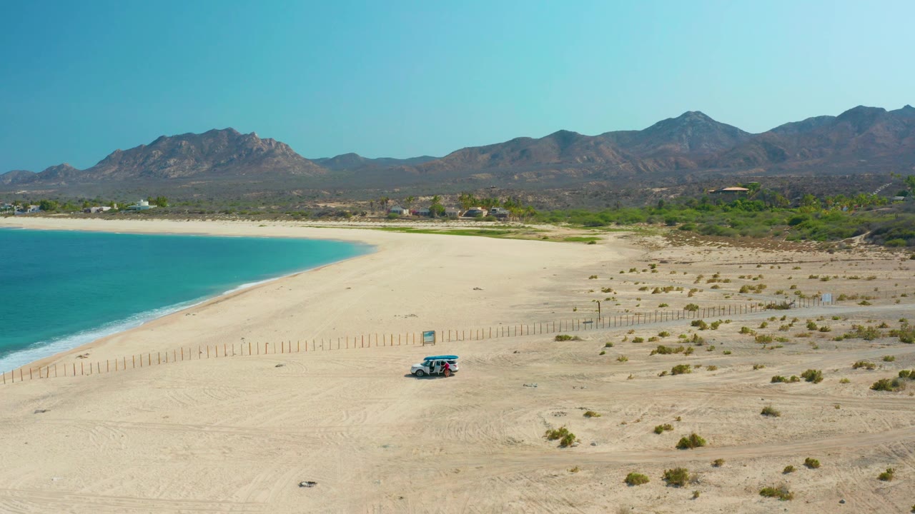 At mid-clip, the small vehicle remains on the sandy Los Frailes beach near the fence line, with blue water at left, mountains across the background, and no visible people.