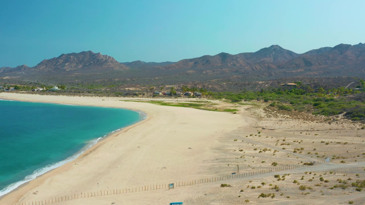 Aerial shoreline view with clear blue water, broad sand, fence line, scrubland, mountains, and vehicle near bottom.